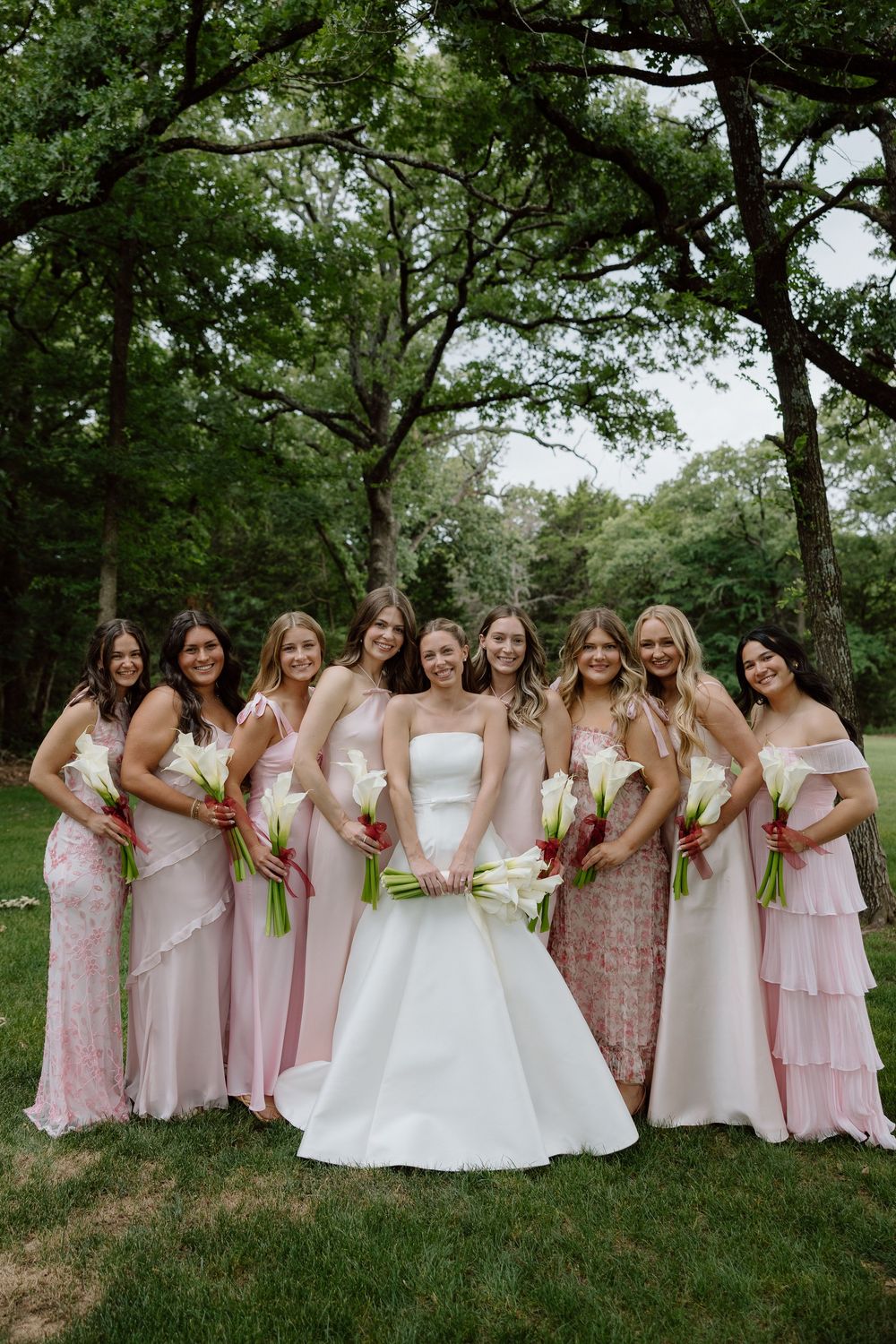 Bride in white gown poses with eight bridesmaids in pink dresses holding white bouquets outdoors.