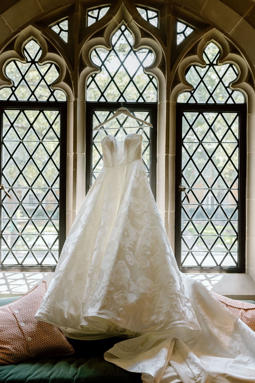A wedding dress hangs elegantly against gothic-style church windows with diamond-patterned leaded glass.