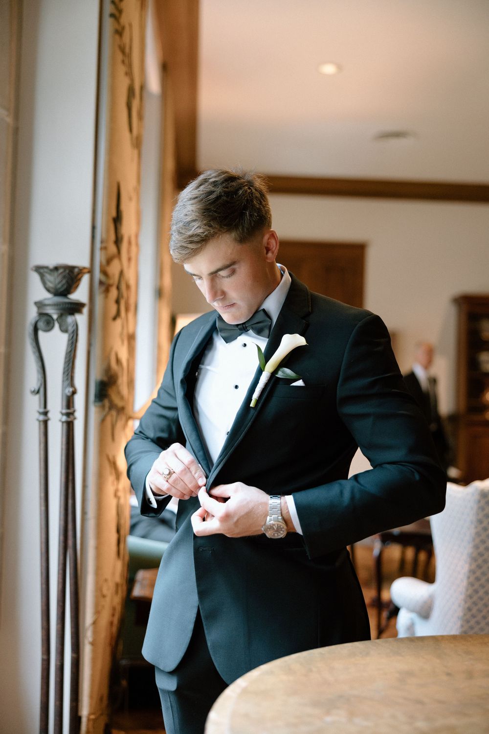 A groom in a black tuxedo checks his watch while getting ready in an elegant hotel room.