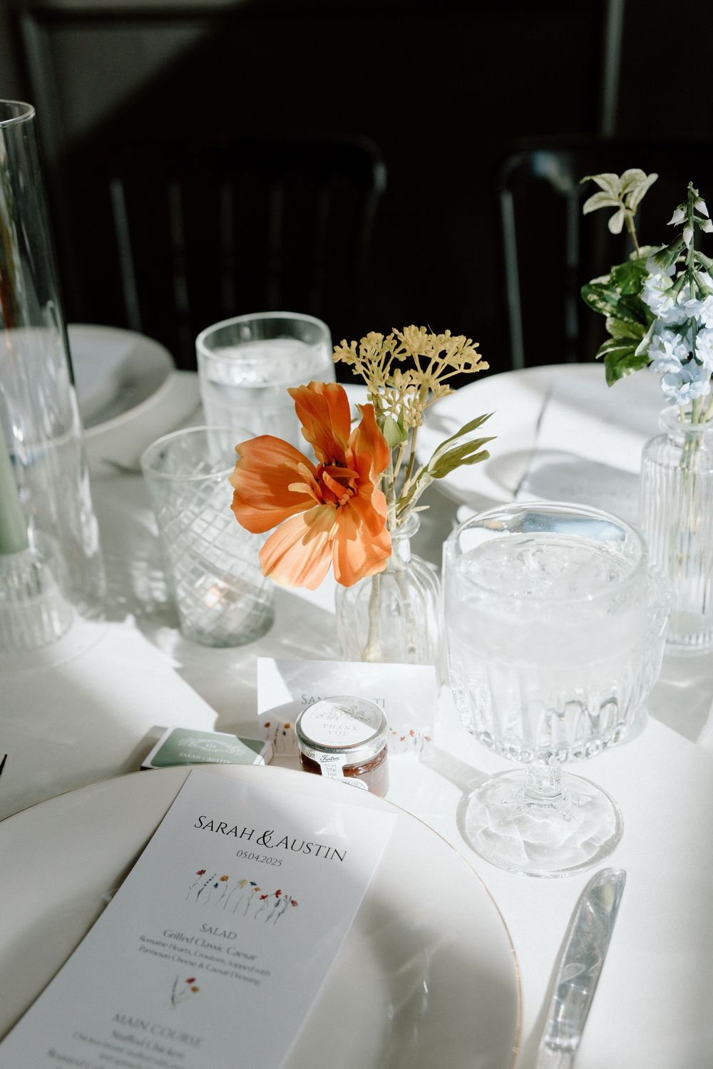 Elegant wedding table setting with orange flower centerpiece, crystal glasses and personalized menu card in natural light.