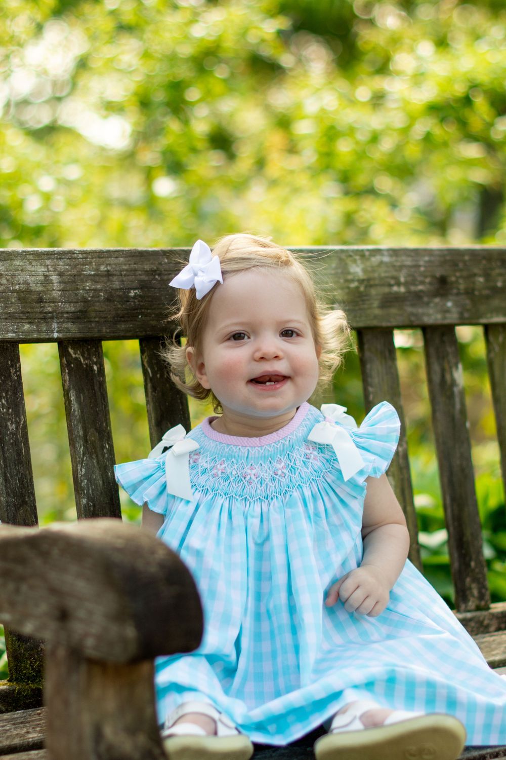 Young child in blue gingham dress sitting on wooden garden bench with bow in hair.