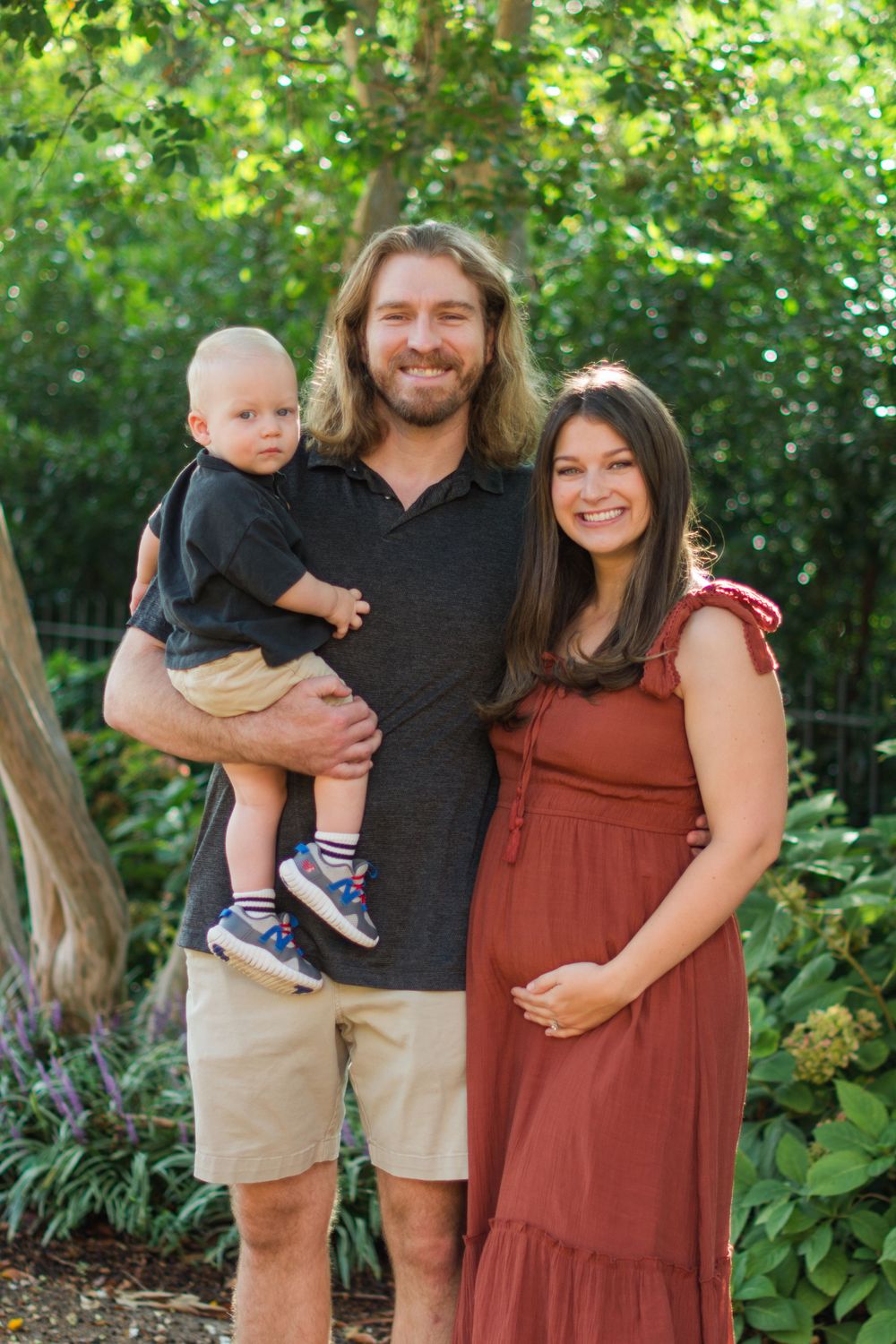 A happy family portrait taken outdoors with a young child and expectant mother in a garden setting.