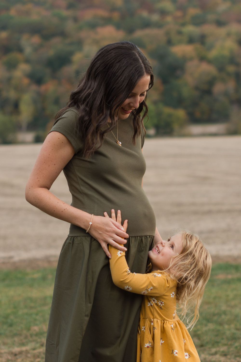 A touching moment between an expectant mother in olive dress and young child in yellow dress outdoors.