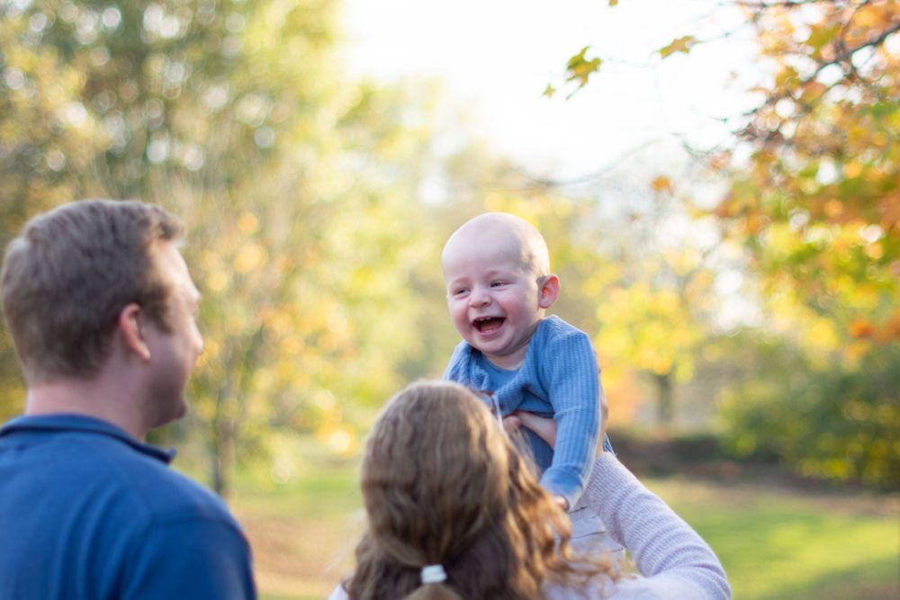 A joyful family moment captured outdoors with a laughing baby lifted high in autumn sunlight.