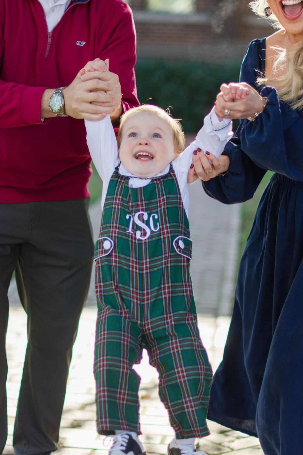 A toddler wearing plaid overalls swings between adults while laughing outdoors.