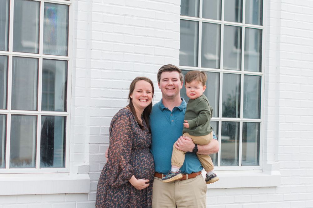 Family of three poses together against white building with large windows.