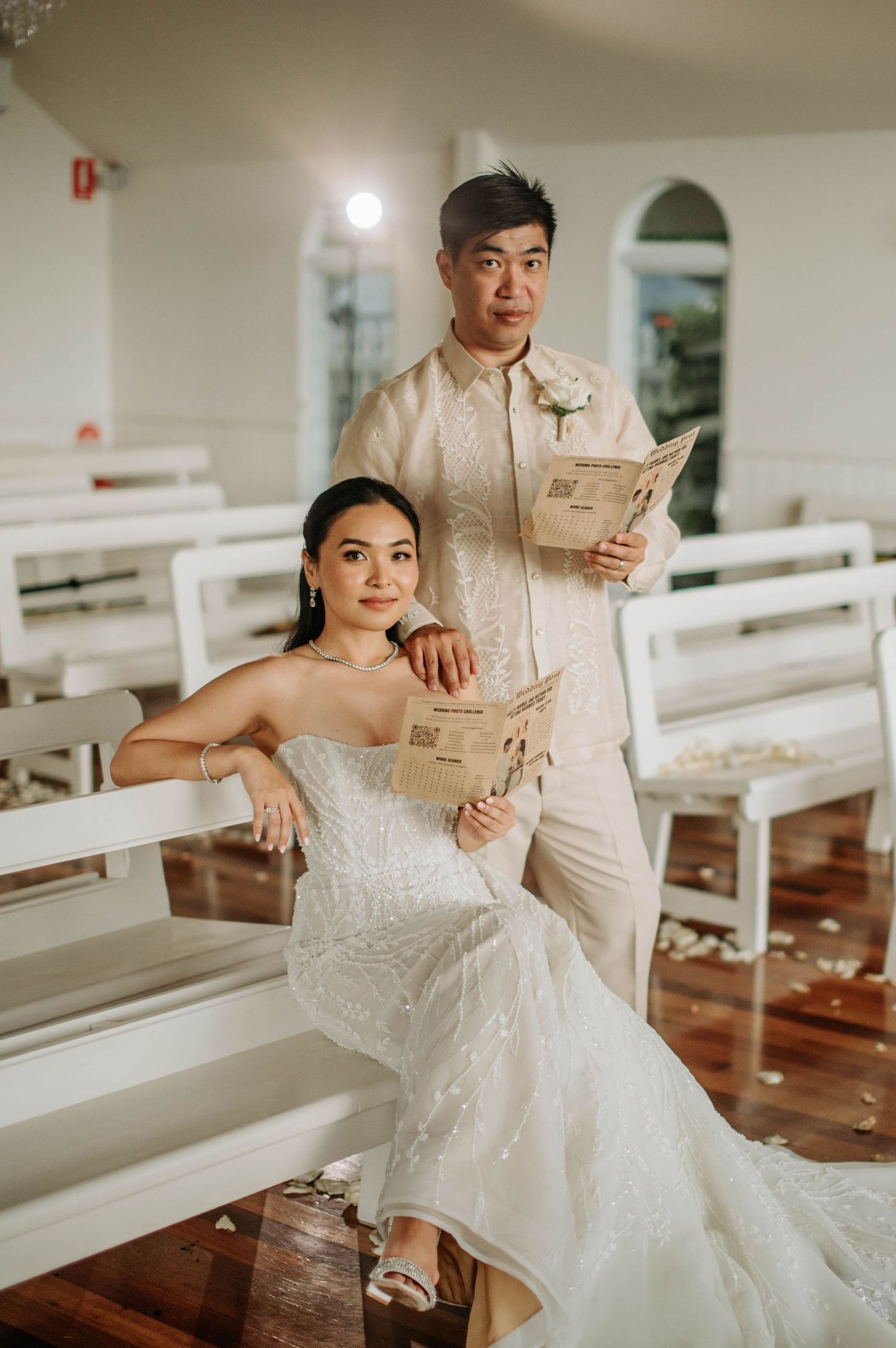 A couple poses casually on white church pews while reading newspapers in wedding attire.