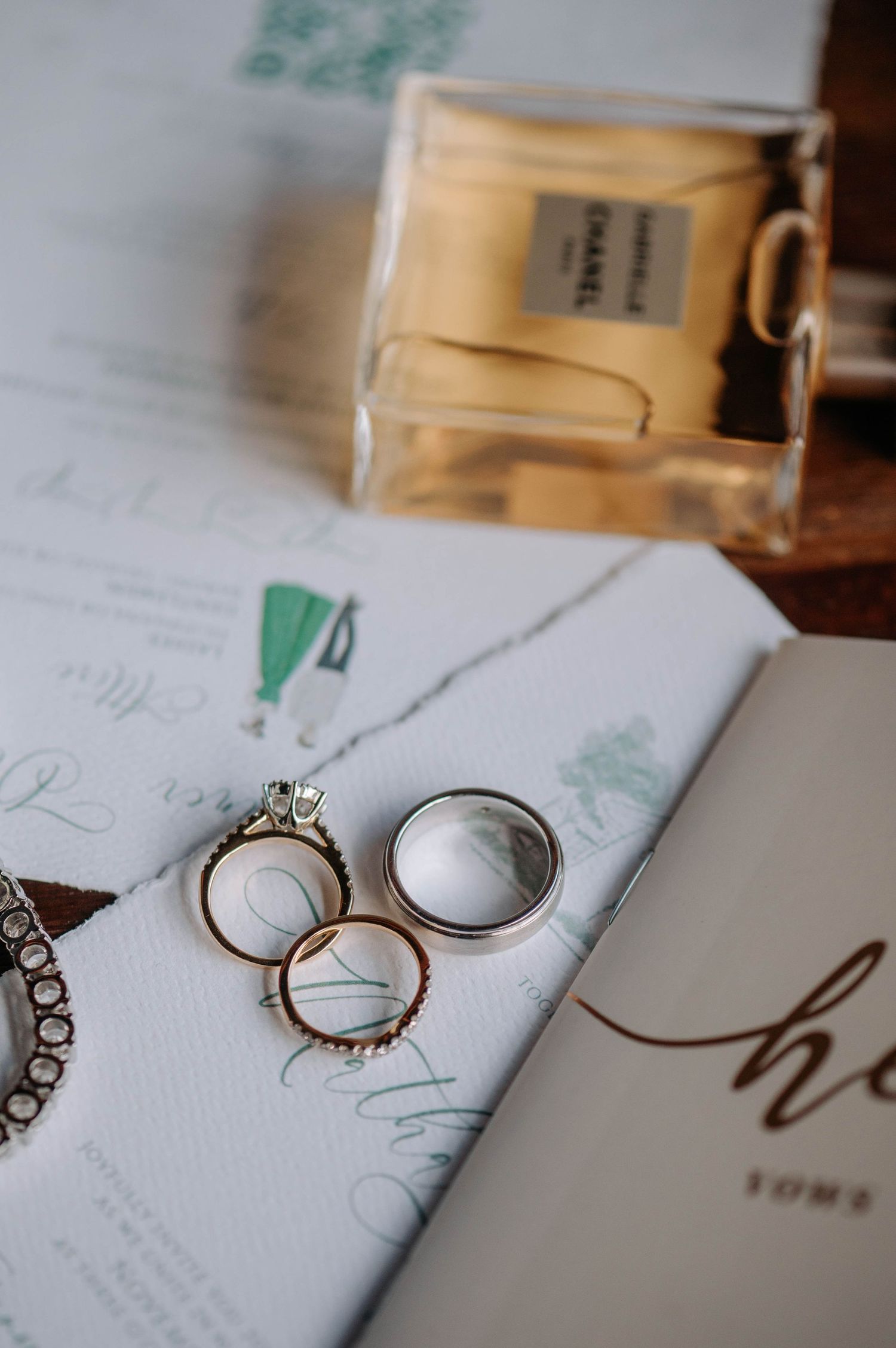 Wedding rings and perfume bottle arranged with invitation stationery on wooden surface.