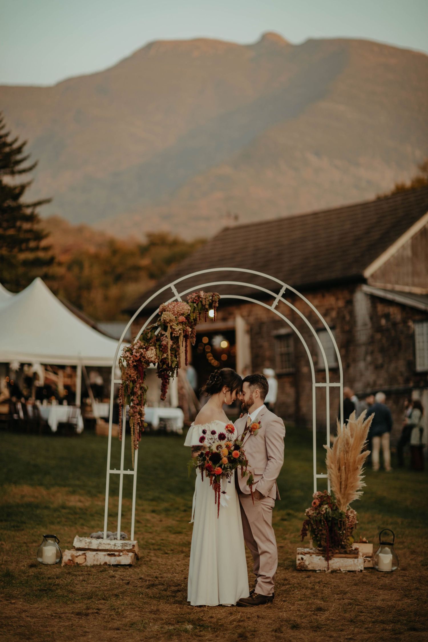 Couple stands under floral wedding arch with mountain backdrop at sunset ceremony.