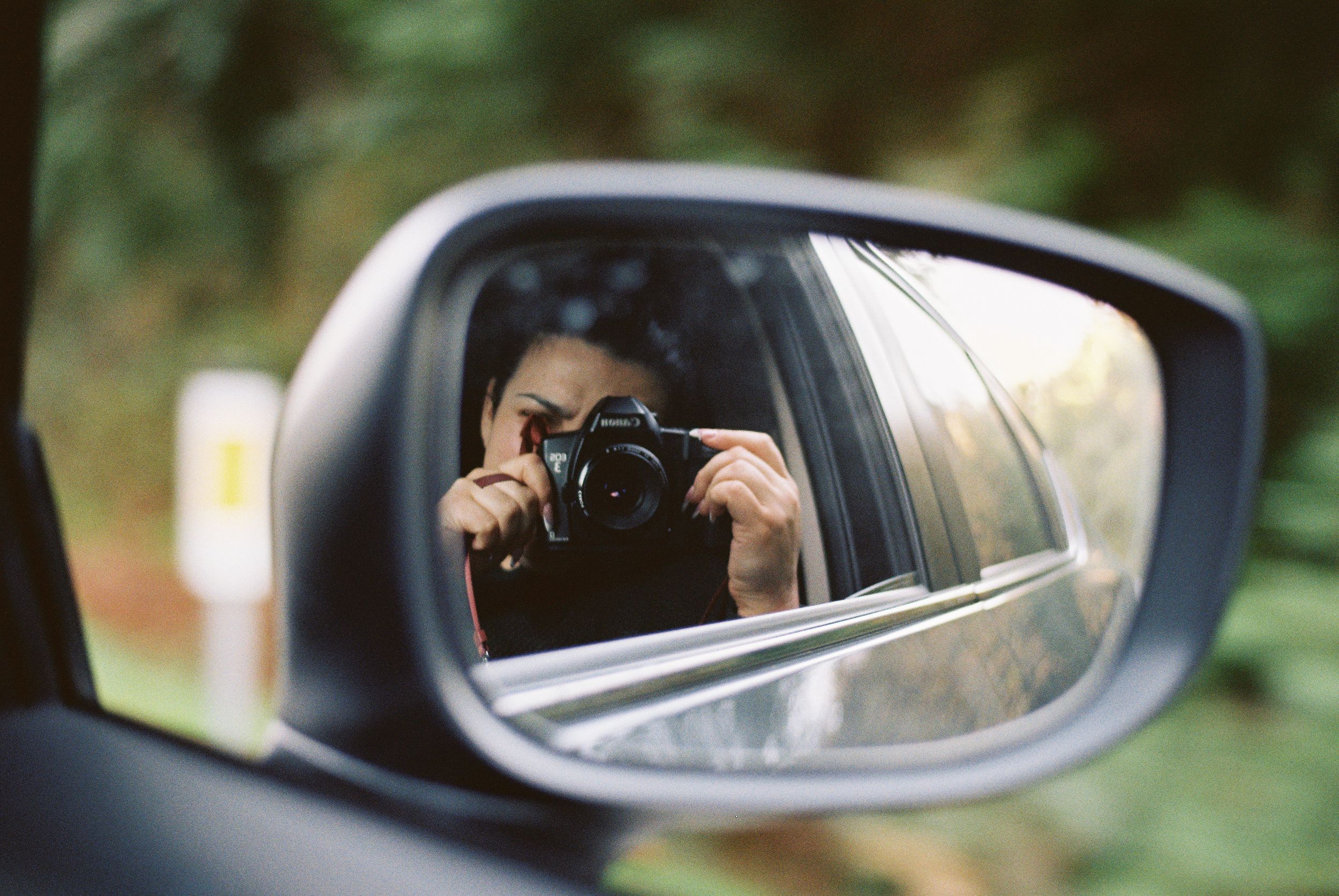Camera reflected in car side mirror capturing a photo against blurred green background.