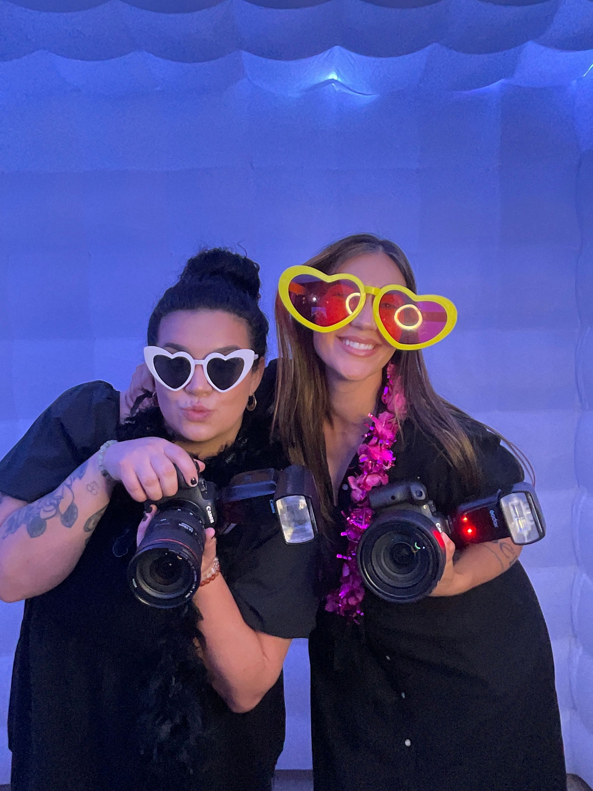 Photographers wearing fun heart glasses pose with cameras against blue backdrop.
