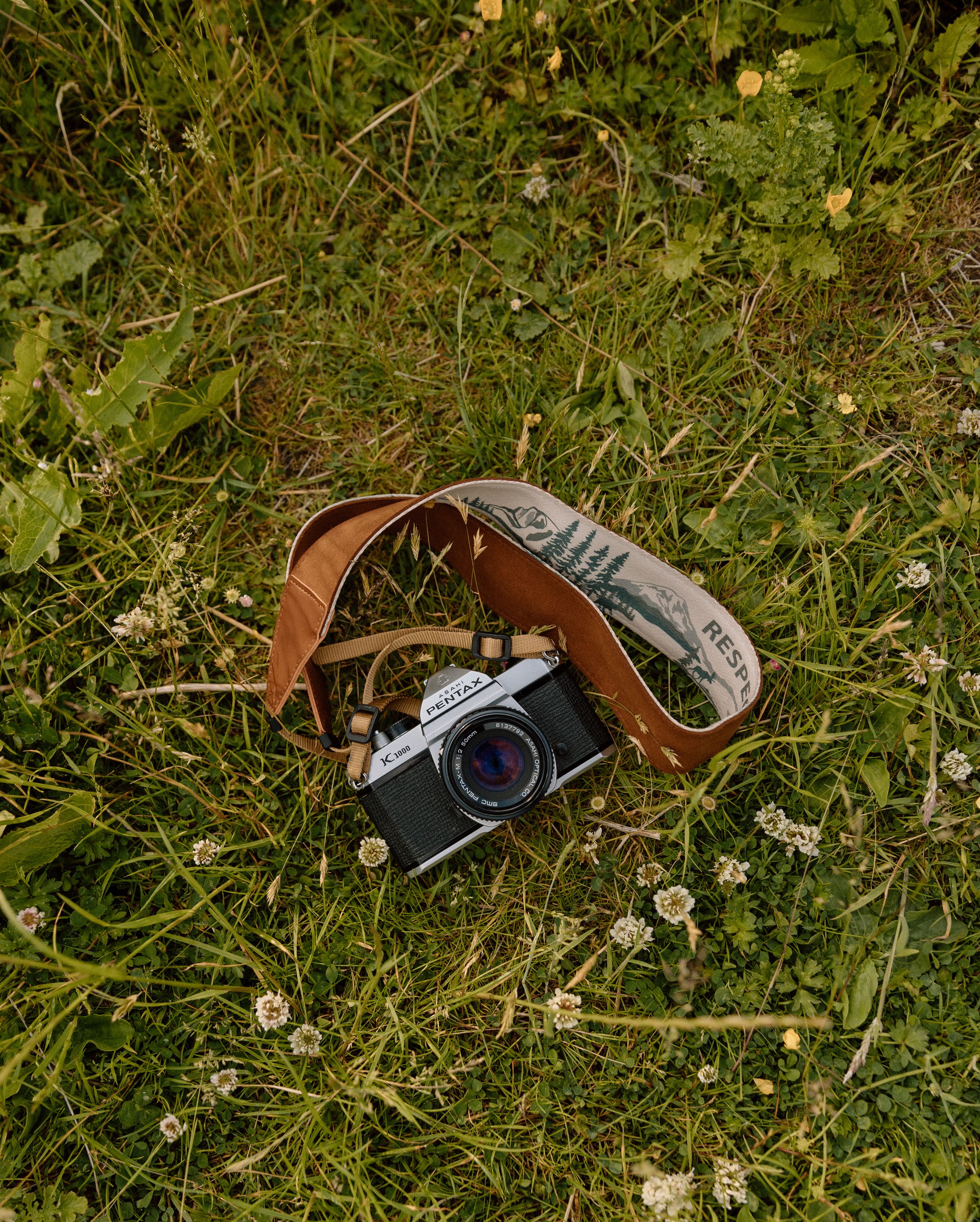 Vintage film camera with brown leather strap lying in grass with small white flowers.