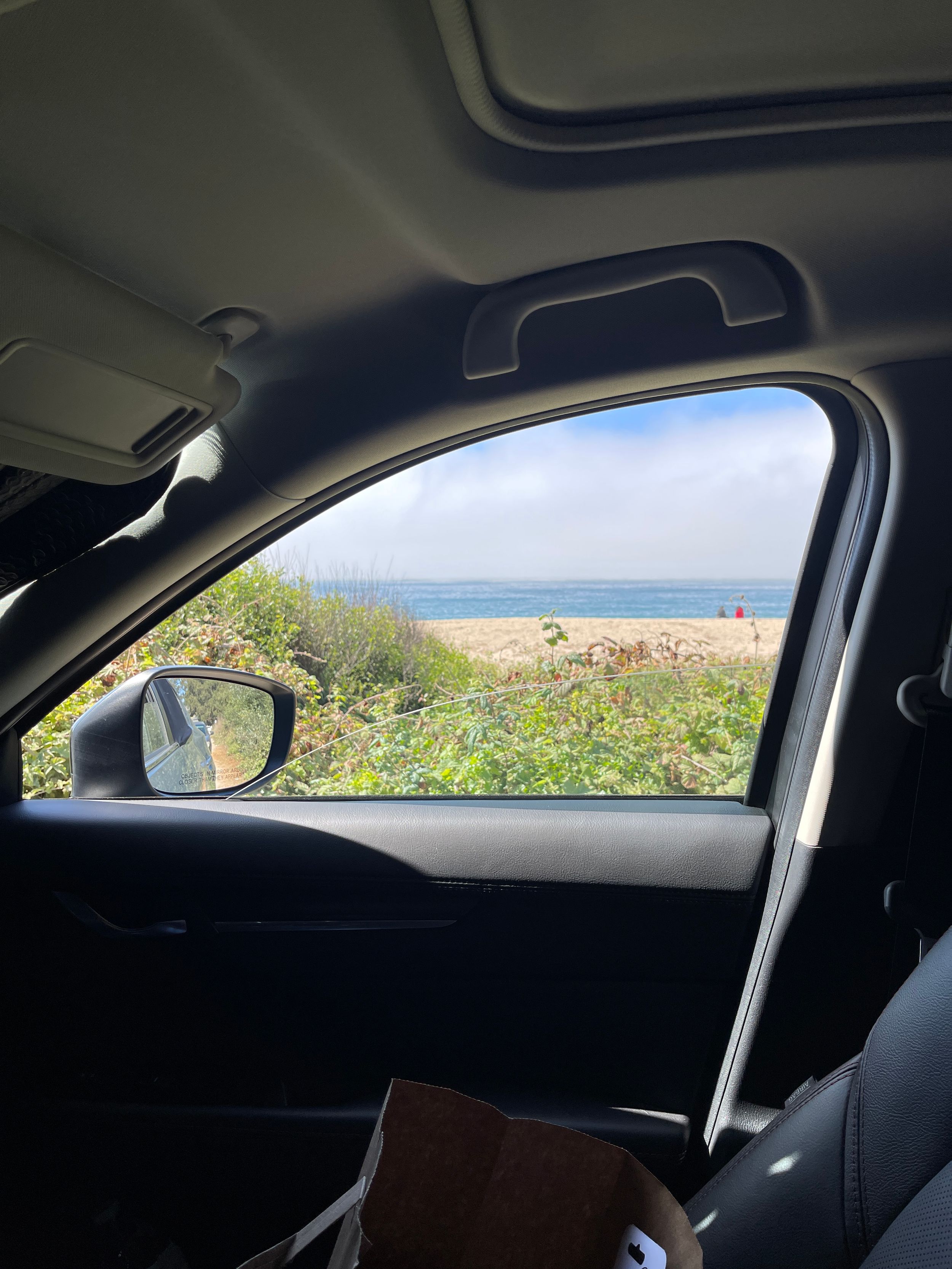 View of a beach and ocean through a car window with side mirror visible.
