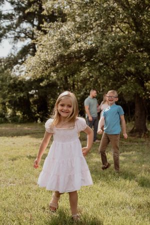 Young girls in white dresses play in a sunlit grassy field during a photo session.