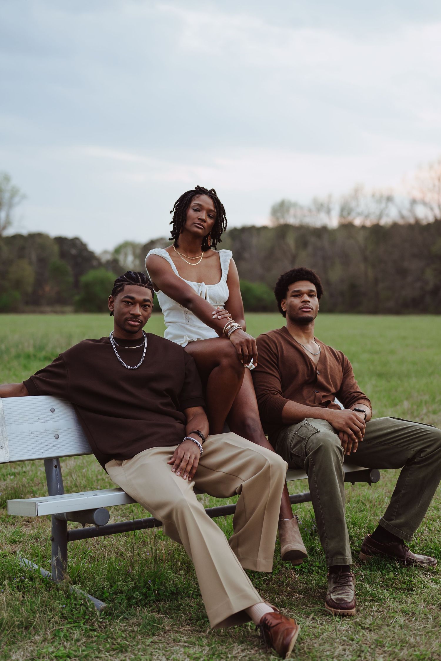Three young adults pose outdoors on a bench in a green field, wearing earth-toned casual outfits on a cloudy day.