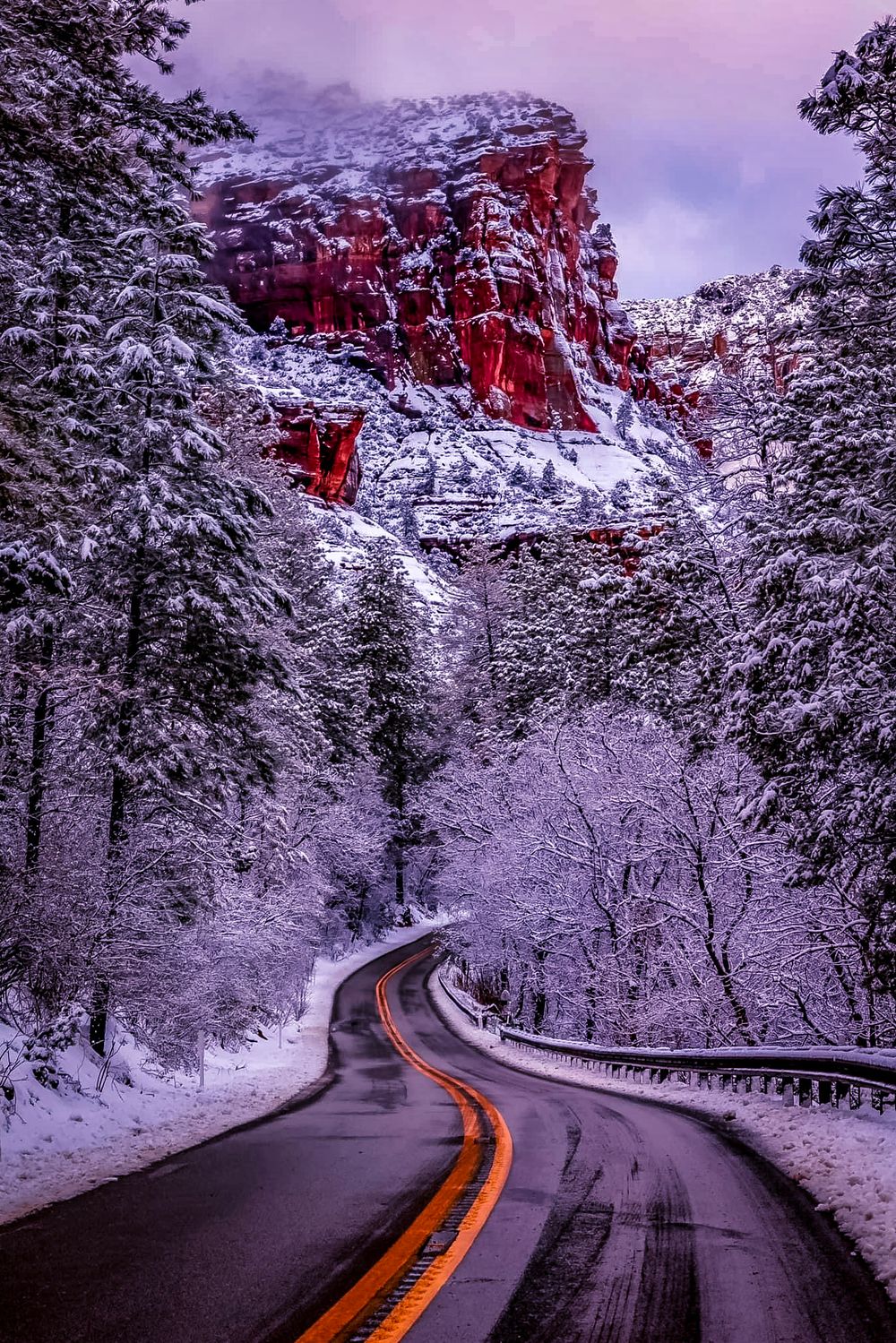 Snowy winter road curves through red rock cliffs in a picturesque mountain landscape.