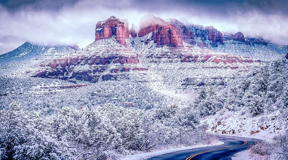 Snow-covered red rock formations in Sedona Arizona with dramatic cloudy skies and winding mountain road.