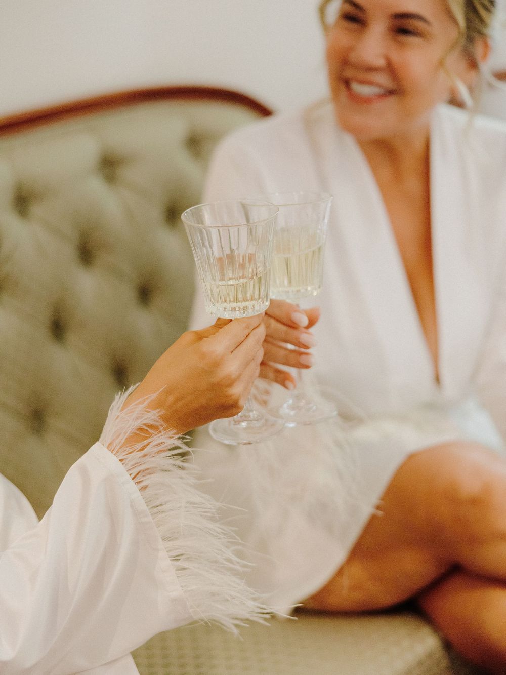 Two champagne glasses toasting during wedding preparations on an elegant tufted sofa.