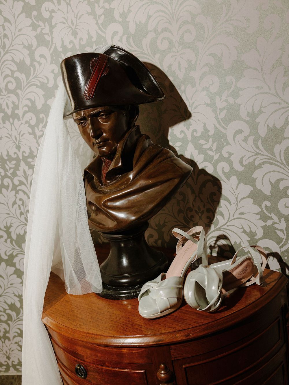Decorative bronze bust with wedding accessories on a wooden table against damask wallpaper.