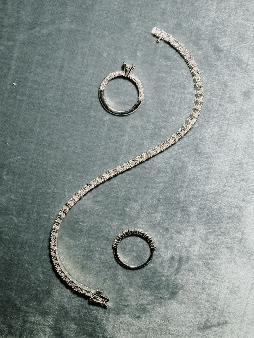 Diamond jewelry arranged in a yin-yang pattern with wedding rings and a tennis bracelet on gray background.