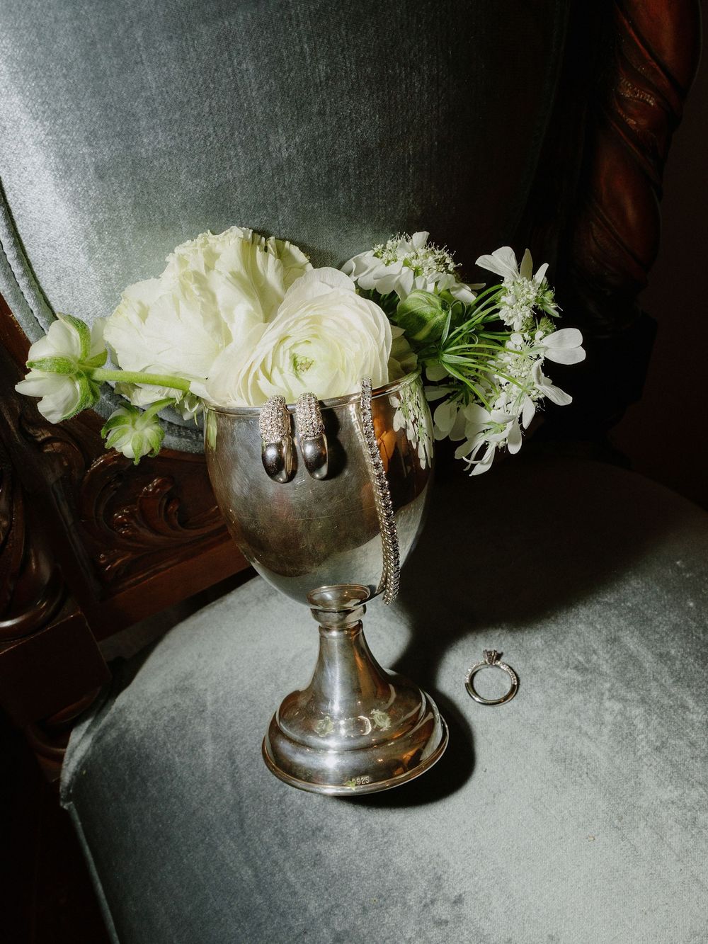 White roses and flowers arranged in a vintage silver goblet with a wedding ring in the background.