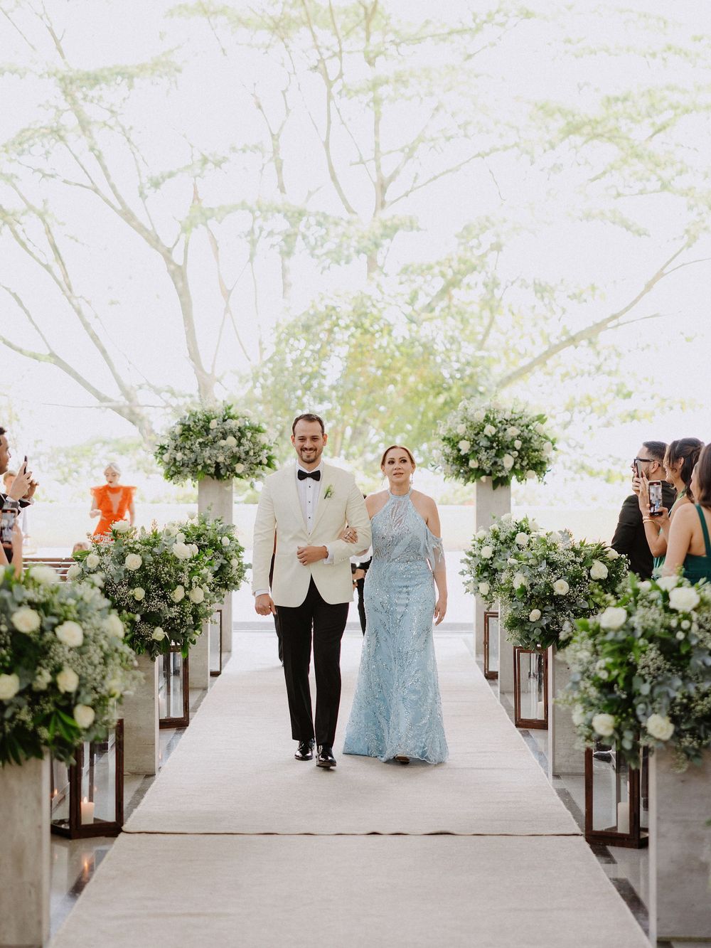 Wedding processional on white carpet with lush green floral arrangements lining the aisle.