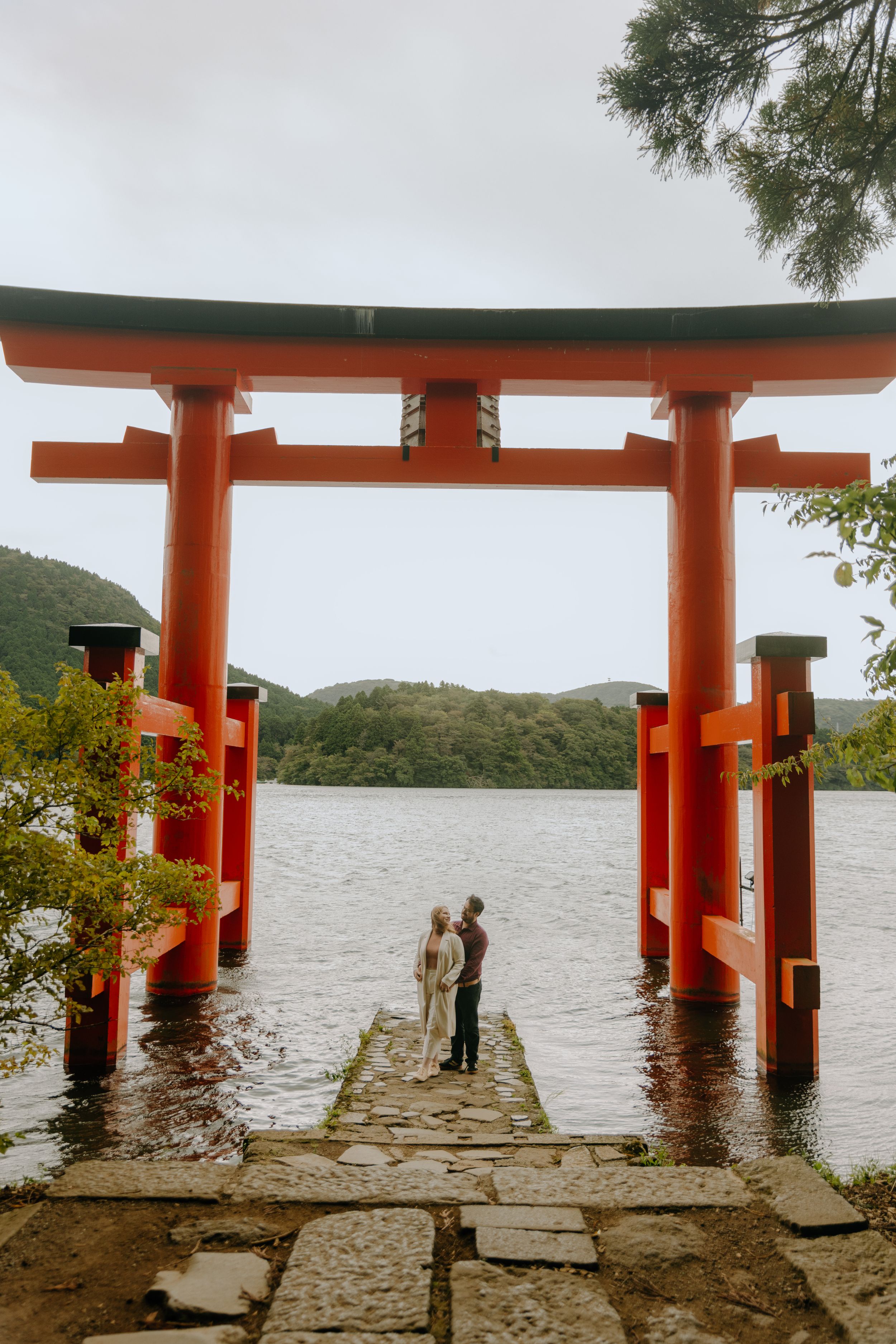 A couple stands beneath a traditional red Torii gate overlooking a serene lake in Japan.