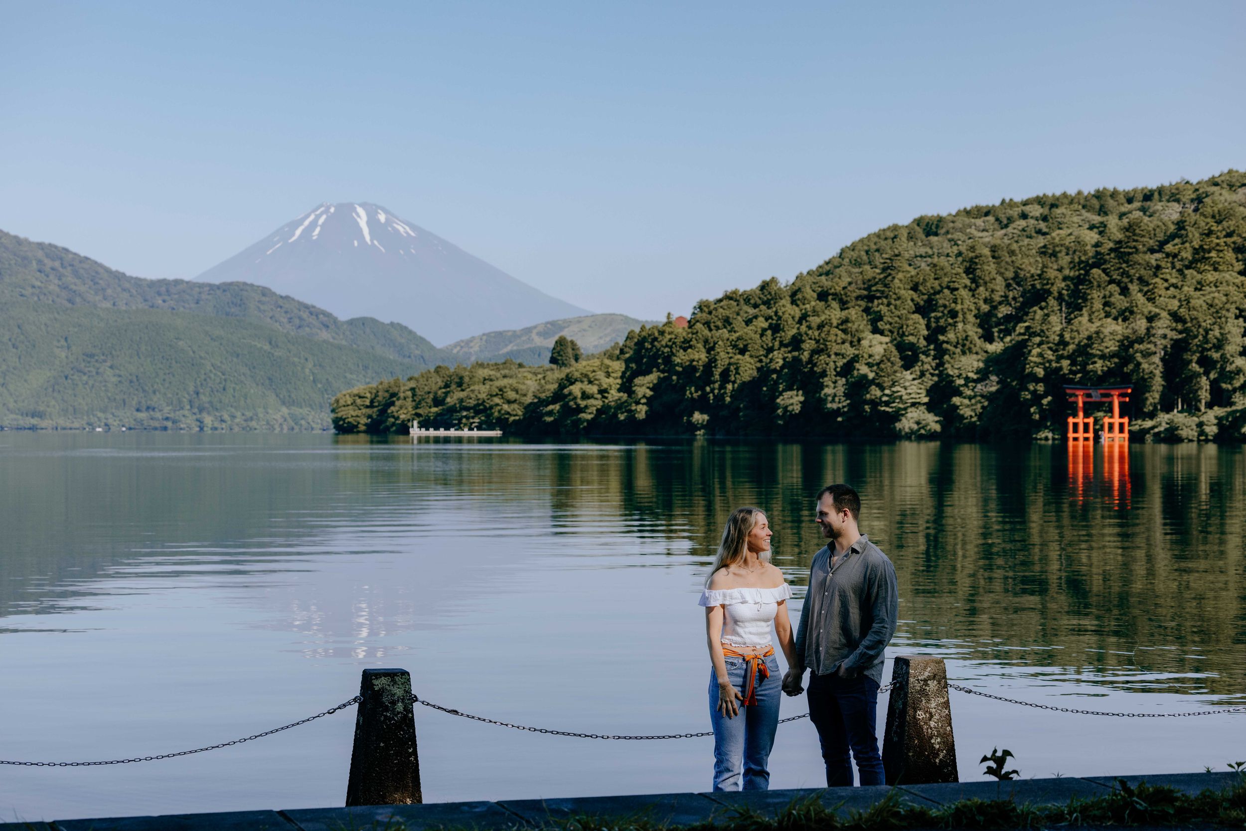 Two people stand by a serene lake with Mount Fuji and a red torii gate visible in the background.
