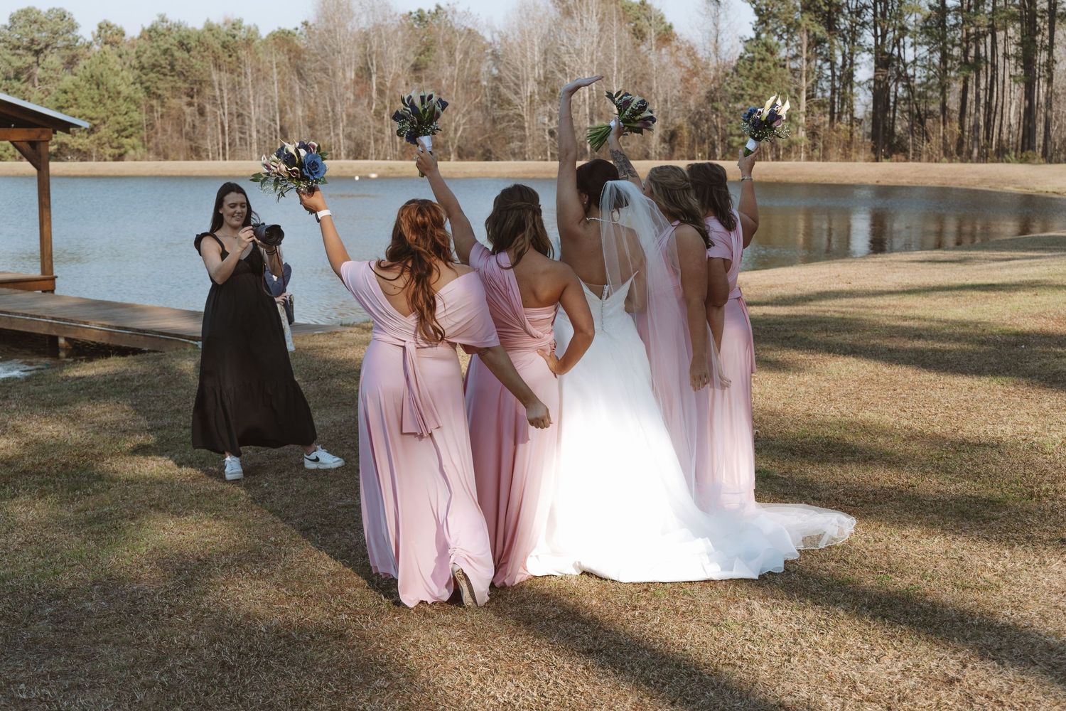 Wedding party poses together by a lake, with bridesmaids in pink dresses raising bouquets.