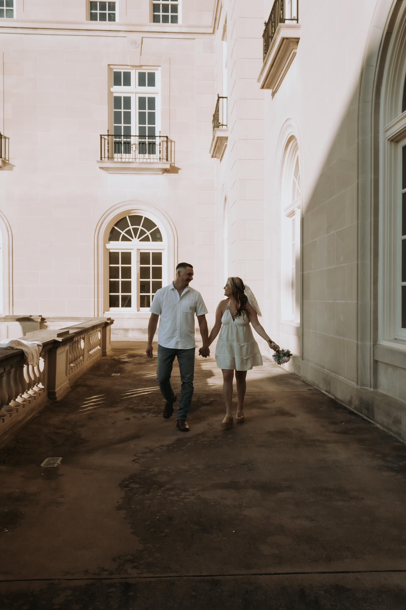 A couple holding hands walks away down a dimly lit corridor in an elegant white building.
