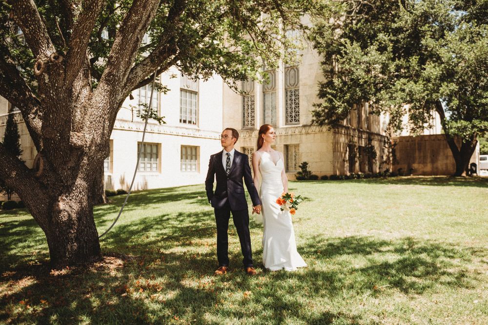 A couple in wedding attire stands together on a sunny lawn beside a large tree near a historic building.
