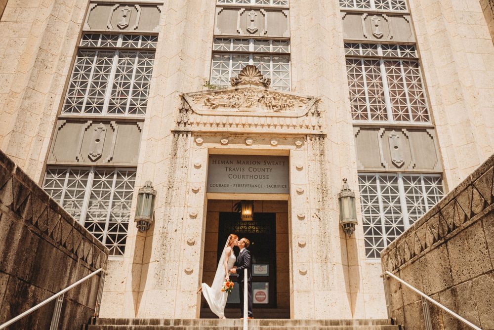 A couple embraces in the grand entryway of an Art Deco-style building with ornate stone details and decorative windows.