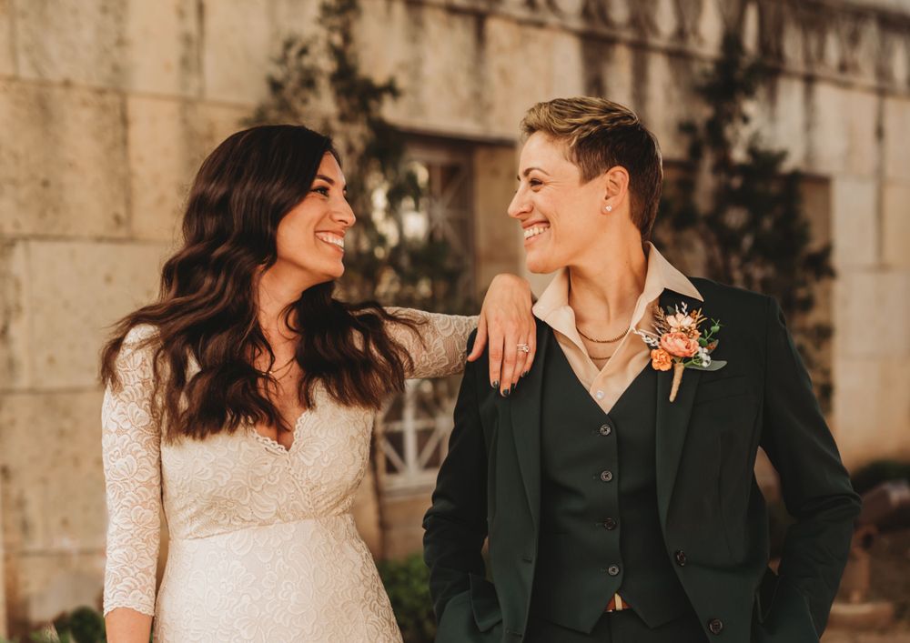 A bride and partner smile lovingly at each other in warm golden light while standing against a stone wall.