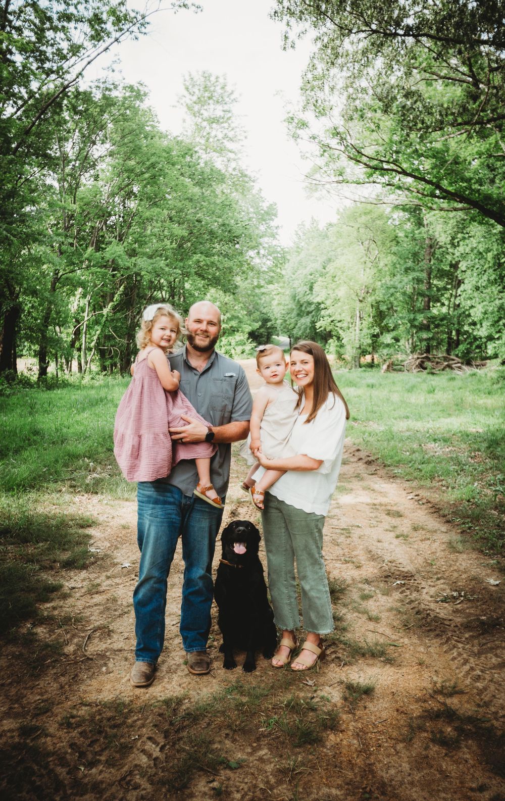 A rural dirt path winds through lush green trees where a family and their black dog gather together on a sunny day.