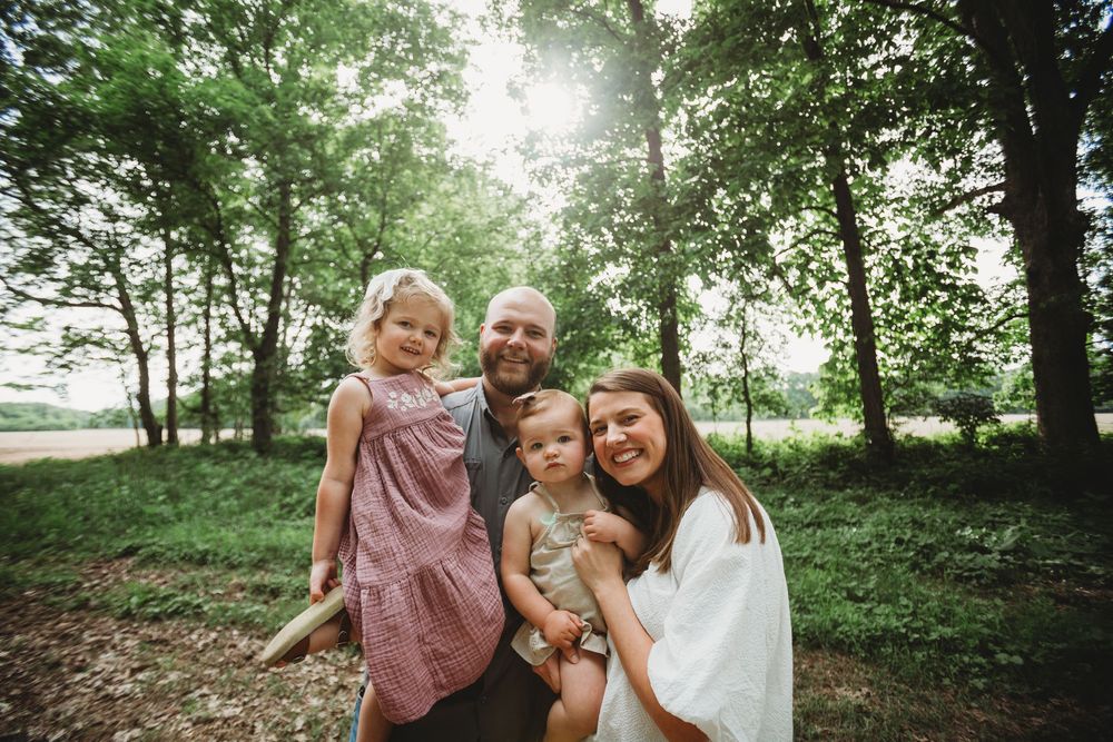 A family poses together in a sun-dappled forest clearing on a bright summer day.