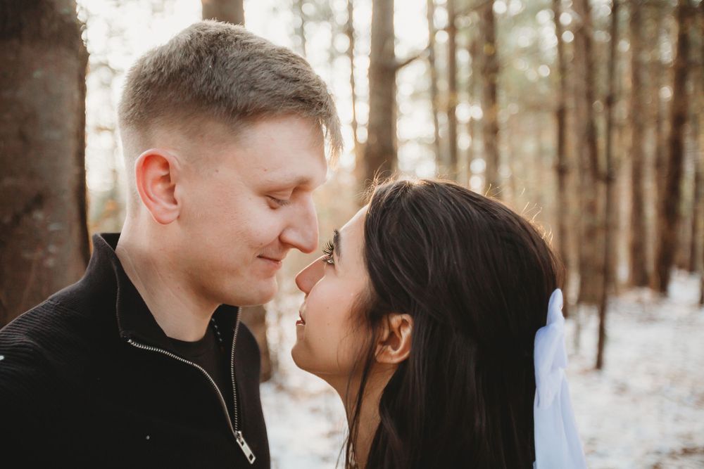 A romantic couple shares an intimate moment in a snowy forest setting with sunlight filtering through the trees.