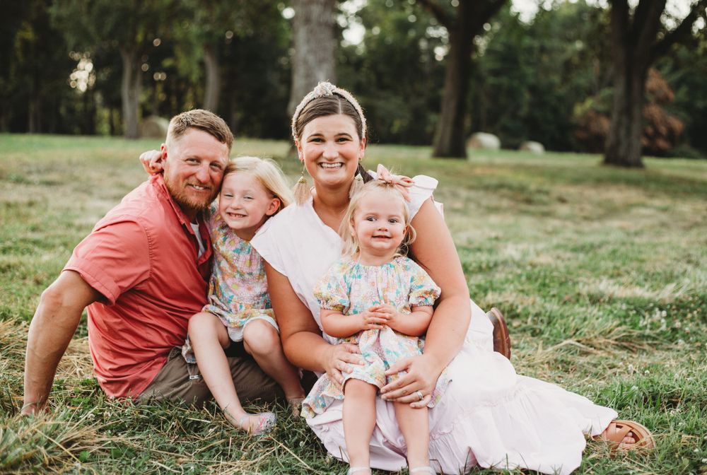 A family of four sits together on grass for a casual outdoor portrait.