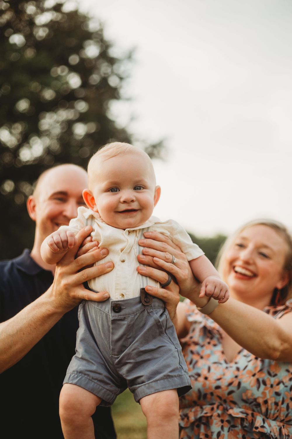 Parents playfully lift their baby wearing grey shorts during a joyful outdoor moment.