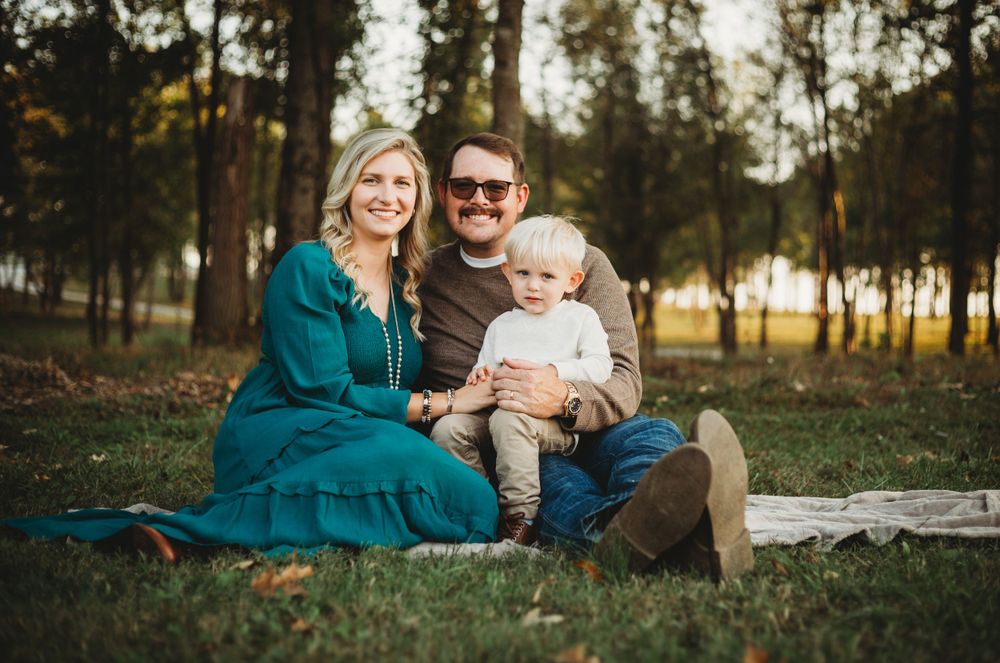 A family sits together on a blanket outdoors during golden hour in a wooded setting.