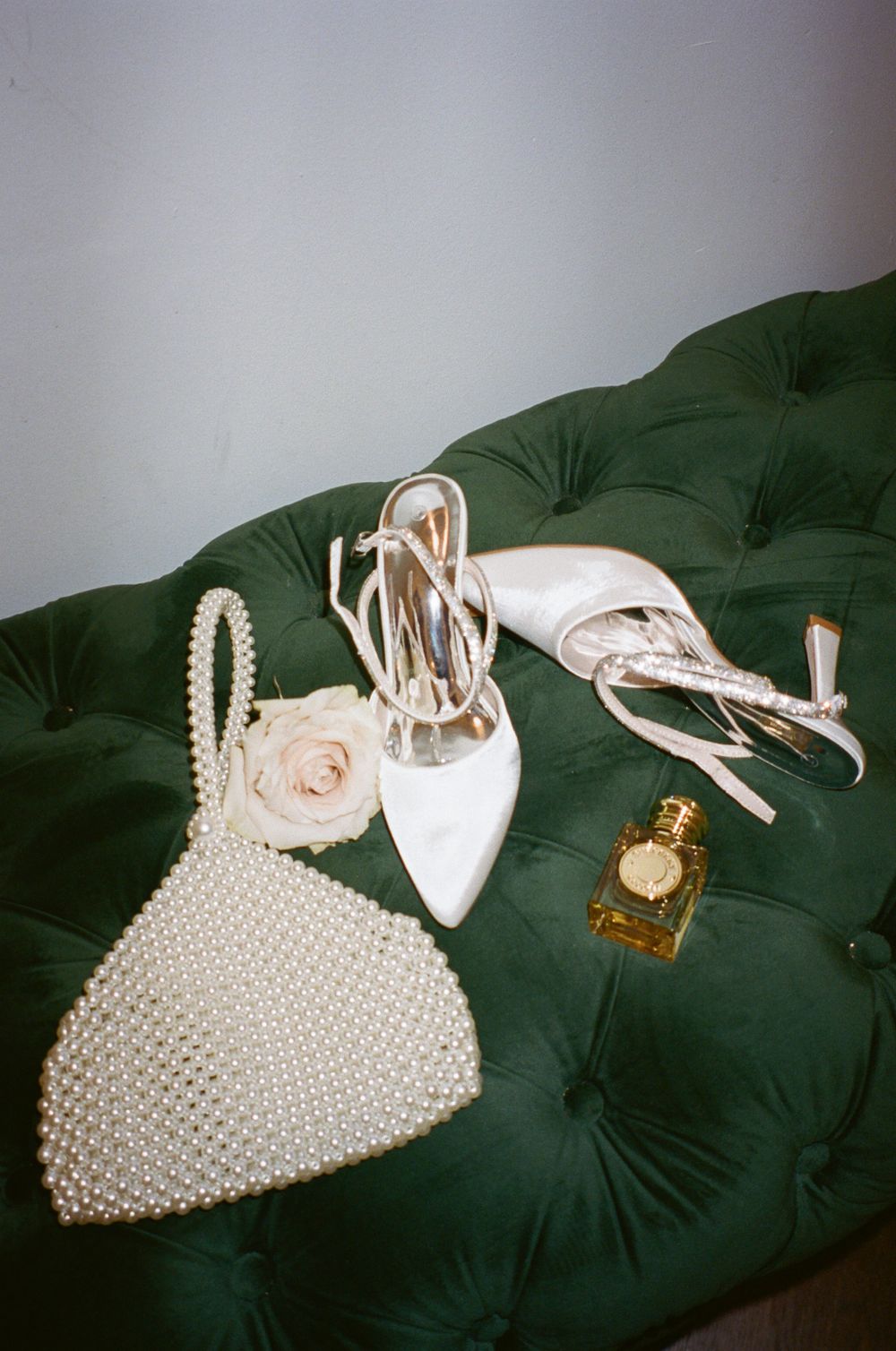 Wedding accessories including white heels, pearl handbag and perfume displayed on green velvet furniture.