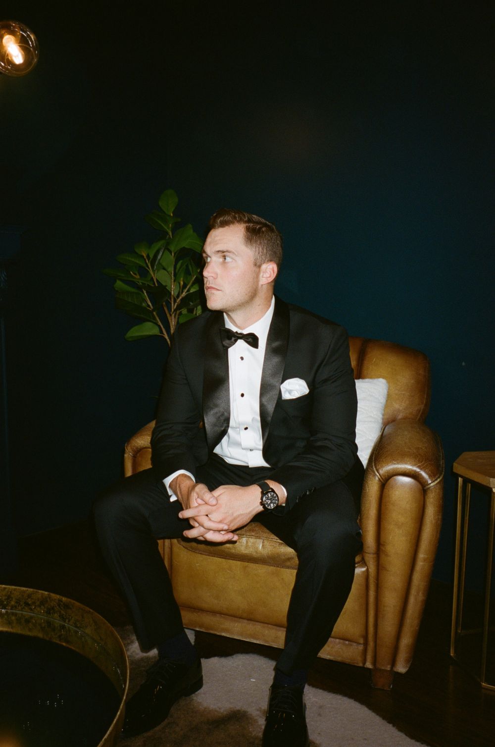 Person in formal black tuxedo seated on leather chair in moody lighting.