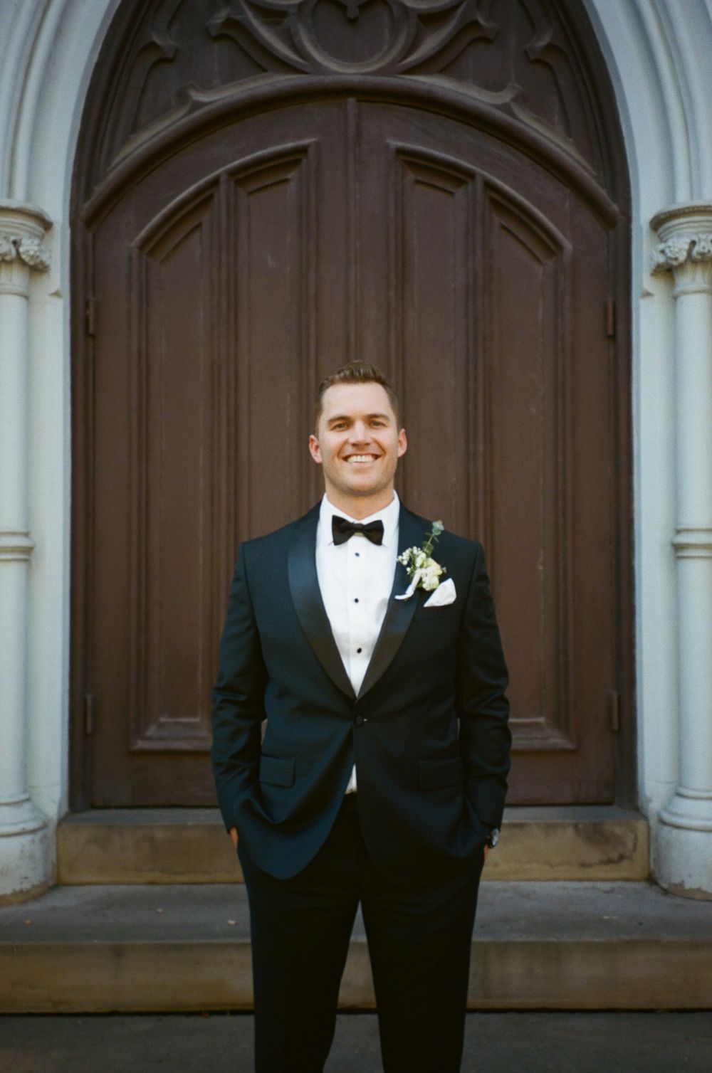 A dapper groom in a black tuxedo and bow tie poses in front of an ornate church doorway.