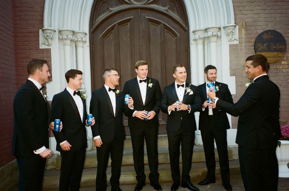A group of groomsmen in black tuxedos gather on church steps sharing drinks and conversation.