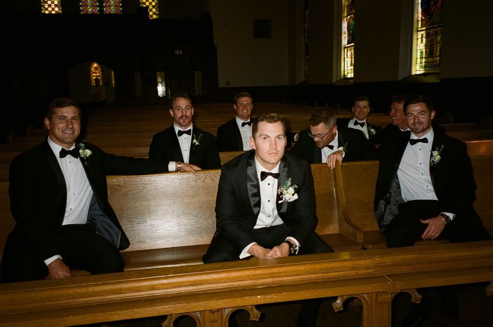 Wedding party members relax in wooden church pews wearing formal black tie attire.