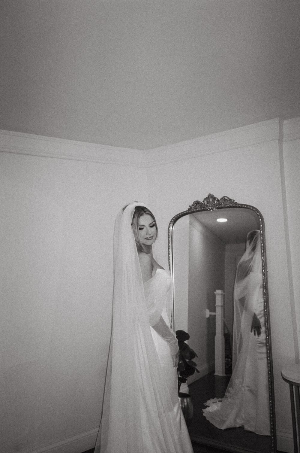 A bride in a white wedding gown looks at her reflection in an ornate mirror during wedding preparations.