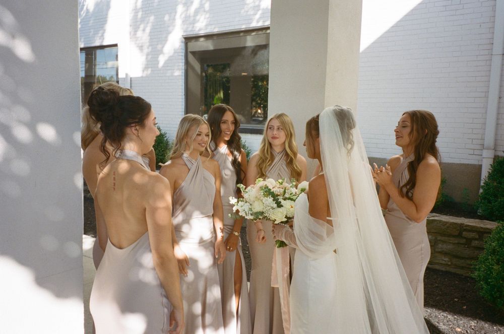 Bridesmaids in light colored dresses surround the bride holding white bouquets in natural sunlight.