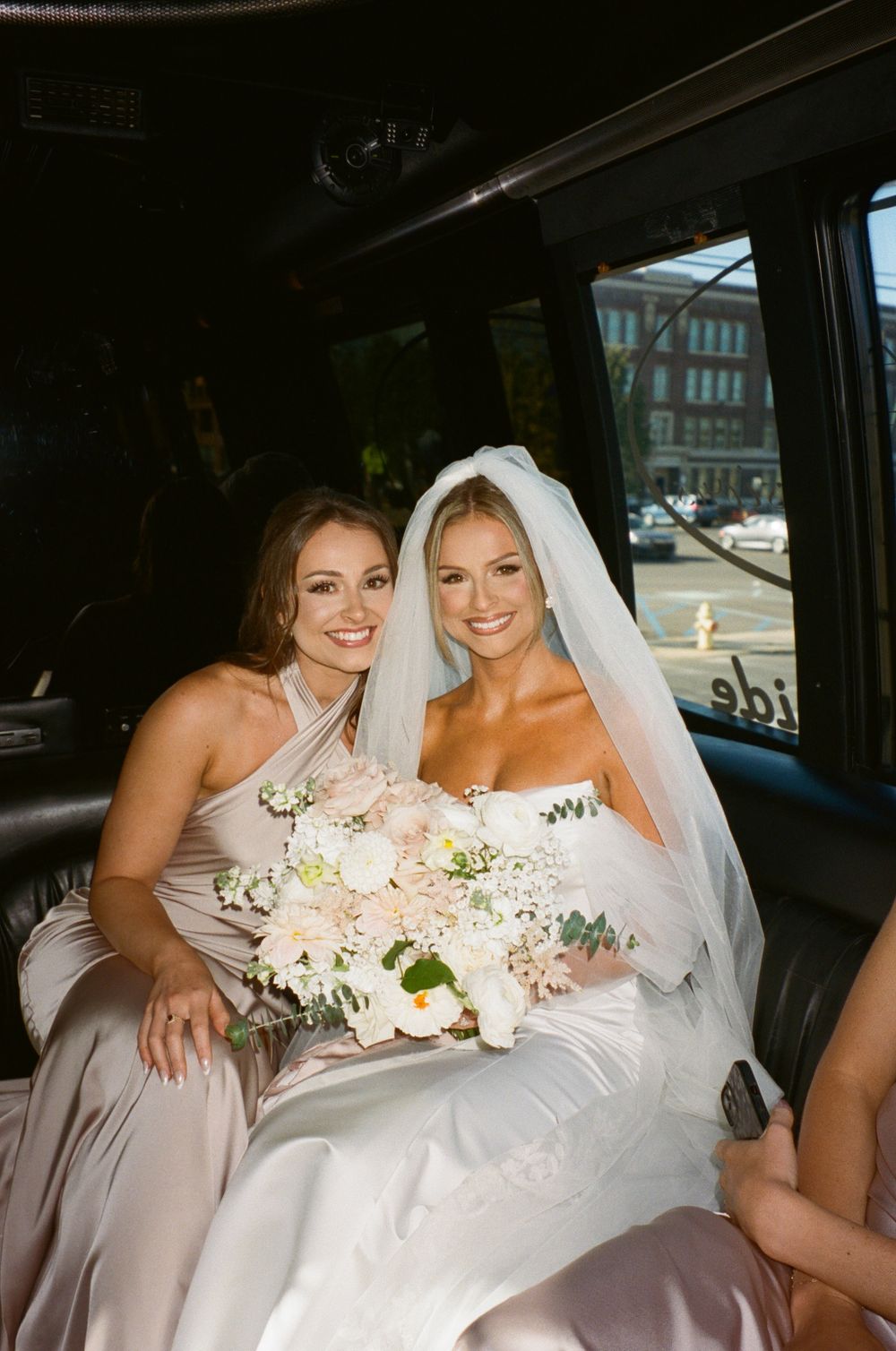 A bridesmaid and bride pose together with bouquets inside a wedding vehicle.