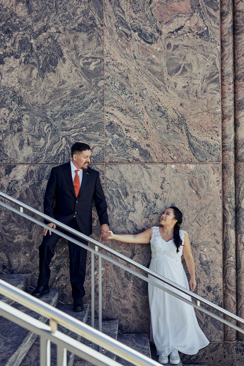 A couple in wedding attire poses on marble stairs with dramatic architectural backdrop.