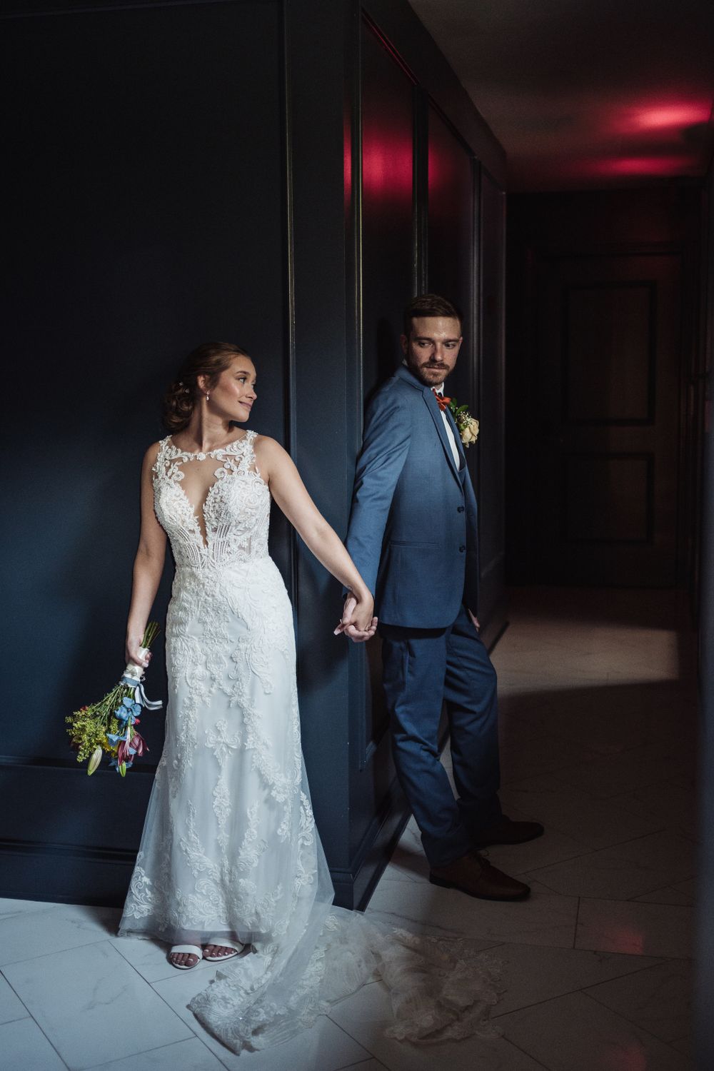 A couple in wedding attire holds hands in a dramatically lit hallway with red ambient lighting.