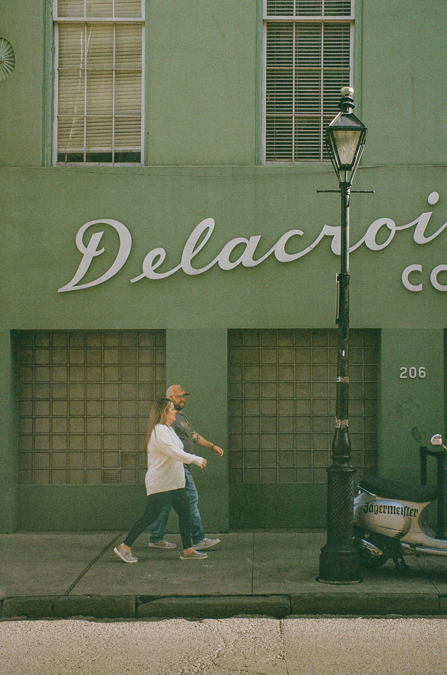 A pedestrian walks past a green building with 'Delacroi' signage and vintage street lamp in an urban setting.